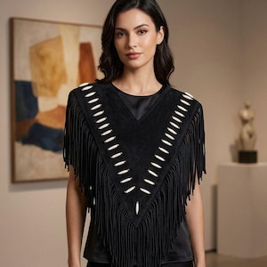 Puede incluir: Un poncho de ante negro con escote en pico y largos flecos en los bordes. La prenda está decorada con dos filas de cuentas cilíndricas blancas dispuestas en forma de V en la parte delantera, sobre una camiseta de satén negro.