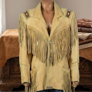 May include: A tan leather jacket featuring long fringe detailing across the chest and lower pockets. The garment includes a notched collar, front button closures, and decorative geometric beadwork patterns on both shoulders.