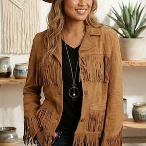 May include: A tan suede jacket featuring long fringe detailing on the chest, sleeves, and hem, paired with a brown felt fedora hat and a long silver-tone necklace with a circular pendant over a black v-neck shirt and dark blue denim jeans.