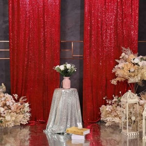 May include: Two floor-length red sequin curtains hang vertically. A small table covered in a silver sequin cloth holds a pink vase with white flowers. A white decorative lantern and floral arrangements are placed on the floor nearby.
