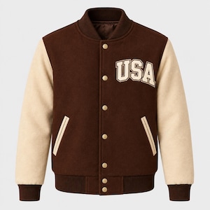 Könnte beinhalten: Eine braune Collegejacke mit cremefarbenen Ärmeln sowie geripptem Kragen, Bündchen und Saum. Auf der Vorderseite befindet sich der Schriftzug USA in cremefarbenen Buchstaben mit dunkelbrauner Umrandung, fünf Druckknöpfen und zwei Seitentaschen.