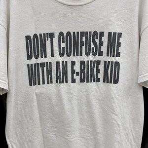 Puede incluir: Una camiseta de manga corta y cuello redondo en color blanco roto con texto en letras negras de bloque impreso en el pecho que dice: "DON'T CONFUSE ME WITH AN E-BIKE KID." La prenda se muestra en una percha de plástico negra.