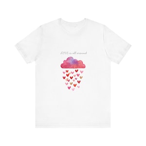 May include: A white short-sleeved t-shirt featuring a watercolour-style graphic of a pink and purple cloud raining small red and pink hearts. Above the cloud, the text reads "LOVE is all around" in a simple, thin, dark-coloured cursive font.