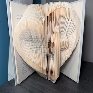 May include: A book with pages folded to create a three-dimensional heart shape. The off-white paper pages are precisely creased and fanned outward to form the central heart design, whilst the book covers remain open at the sides to support the structure.