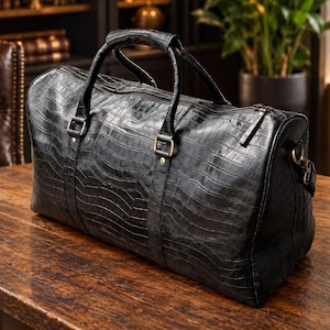 Peut inclure: Un sac de voyage noir avec un motif texturé en peau de crocodile. Le sac présente deux poignées supérieures, une fermeture éclair principale et des détails en métal. Il possède une base rectangulaire et une forme structurée.