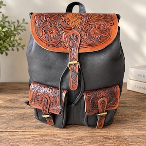Può includere: Uno zaino in pelle nera con patta e due tasche frontali in pelle color cuoio, decorate con intricati motivi floreali incisi. Lo zaino presenta una cinghia centrale verticale con fibbia in ottone e fibbie abbinate sulle tasche anteriori.
