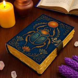 May include: A rectangular journal with a dark blue cover featuring a gold and orange scarab beetle illustration, floral motifs, and a sun symbol. The book has a metal latch closure and thick, textured, yellow-toned paper edges.