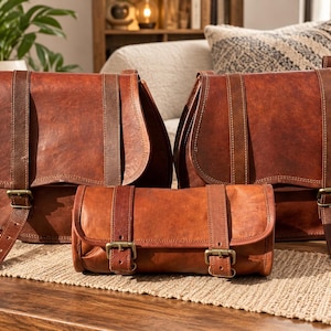 May include: Two large brown leather saddle bags with rounded flaps and vertical straps, positioned behind a smaller cylindrical brown leather roll bag. Each bag features antique brass-toned metal buckles and visible stitching along the edges.