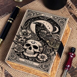 May include: A rectangular journal with a cream-colored cover featuring a black ink illustration of a raven perched on a human skull surrounded by flowers and a crescent moon. The word DEATH is printed at the top and bottom. It has a metal latch.