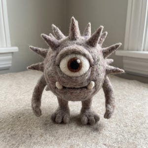 May include: A needle-felted creature with a round, light brown body covered in soft spikes. It features a single large eye with a brown iris and a white sclera, two small white tusks protruding from its mouth, and two short arms and legs.