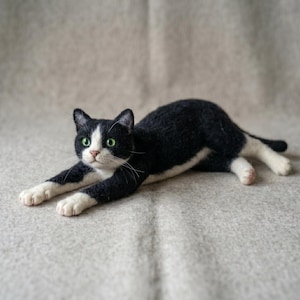 May include: A needle-felted cat figurine with black and white fur, featuring bright green eyes, a pink nose, and long white whiskers. The cat is depicted in a reclining, stretched-out pose with its front paws extended forward and white paws.