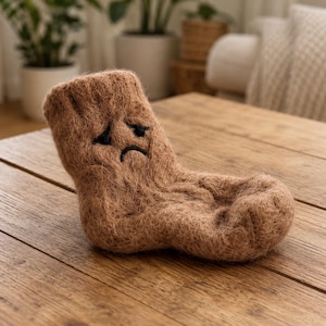 May include: A brown, fuzzy, knitted sock with a sad facial expression embroidered in black thread. The sock has a textured, fibrous appearance and is shaped like a foot, featuring a distinct heel and toe area with a downturned mouth and eyes.