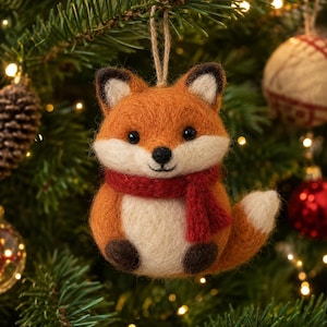 May include: A needle-felted fox ornament featuring orange and cream-colored wool, wearing a small red scarf. The fox has dark brown ears, paws, and eyes, with a white-tipped tail. A jute twine loop is attached to the top for hanging purposes.