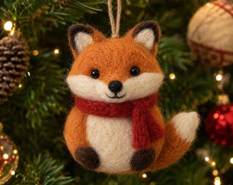 May include: A needle-felted fox ornament featuring orange and cream-colored wool, wearing a small red scarf. The fox has dark brown ears, paws, and eyes, with a white-tipped tail. A jute twine loop is attached to the top for hanging purposes.