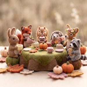 May include: A collection of needle-felted animal figurines including a rabbit, fox, squirrel, hedgehog, deer, and raccoon gathered around a green felted table. The scene features miniature tea cups, plates, a cake, and small orange pumpkins.