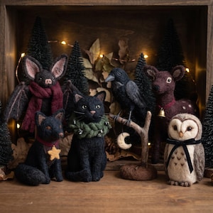 May include: A collection of needle-felted animal figures including a bat with a maroon scarf, a black cat with a green wreath, a black fox with a yellow star charm, a black crow on a branch with a moon charm, a deer with a bell, and an owl.