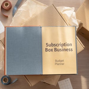 May include: An open book with a blue fabric cover and a tan page featuring the dark brown text Subscription Box Business Budget Planner. The book rests on top of several cardboard shipping boxes with crumpled packing paper inside.