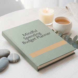 May include: A sage green hardcover book titled Mindful Spending Budget Planner with a horizontal tan stripe across the cover. The book is positioned next to a lit white pillar candle, a small speckled ceramic cup of tea, and smooth gray stones.