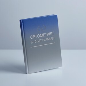 May include: A hardcover planner with a blue to silver gradient cover. The front features a white line-art icon of eyeglasses above the text OPTOMETRIST BUDGET PLANNER, followed by a horizontal line. The book is positioned at an angle.