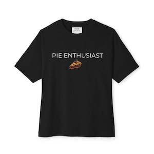 Könnte beinhalten: Ein schwarzes T-Shirt mit kurzen Ärmeln und dem weißen Schriftzug PIE ENTHUSIAST auf der Brust. Unter dem Text befindet sich eine kleine, detaillierte Grafik eines einzelnen Stücks Kirschkuchen mit goldbrauner Kruste und dunkelroter Füllung.