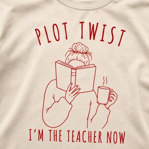 Può includere: Una felpa beige con un disegno a linee rosse di una persona con uno chignon che legge un libro aperto tenendo in mano una tazza fumante. Il testo "PLOT TWIST" è stampato sopra l'illustrazione e "I'M THE TEACHER NOW" è stampato sotto.