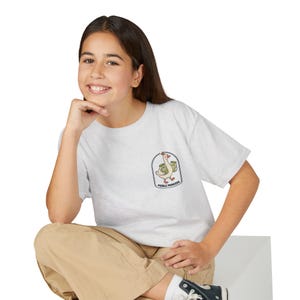Puede incluir: Una camiseta gris claro de manga corta con un pequeño gráfico en el pecho izquierdo. El gráfico muestra un ganso blanco con una pequeña corona que lleva dos frascos de pepinillos, dentro de un arco, con el texto PICKLE PRINCESS debajo.