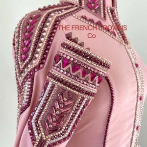Könnte beinhalten: Ein hellrosa Langarmshirt mit aufwendigen Verzierungen aus dunkelrosa und klaren Kristallen, Nieten und metallischen Besätzen an Kragen, Schultern und Ärmeln. Der Text THE FRENCH CROWNS Co ist im oberen Brustbereich aufgedruckt.