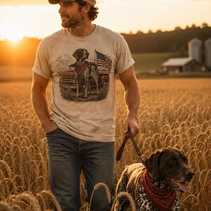 May include: A heathered beige short-sleeve t-shirt featuring a graphic print of a spotted dog wearing a red bandana, standing in front of a red pickup truck and an American flag. The shirt has a crew neckline and rolled sleeve cuffs.