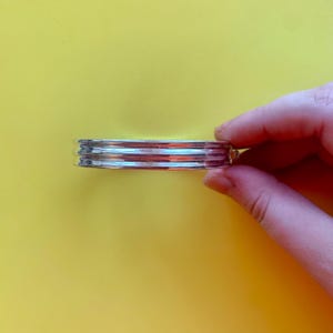 Peut inclure: Un bracelet jonc argenté avec un motif côtelé. Le bracelet est tenu sur un fond jaune. Le bracelet semble être en métal et a une finition brillante. Le design comporte trois arêtes parallèles.