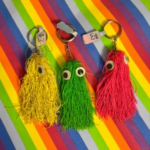 May include: Three handmade keychains in yellow, green, and pink, each featuring a shaggy, wire-like body and large googly eyes. The keychains hang from silver key rings. Price tags are attached to the keychains.