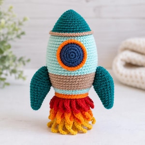 Op de afbeelding: Een gehaakt raket-speeltje met een blauwgroene neuskegel, een lichtblauw lichaam met een donkerblauw en oranje rond venster, en twee blauwgroene zijvinnen. De basis van de raket is versierd met gelaagde rode, oranje en gele garenvlammen.