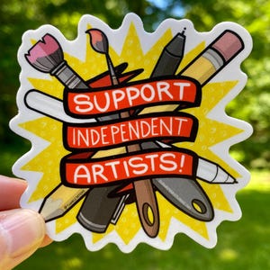 Puede incluir: Una pegatina con un dise&ntilde;o de explosi&oacute;n amarilla que presenta materiales de arte como pinceles, bol&iacute;grafos y l&aacute;pices. Una pancarta roja en el centro dice "SUPPORT INDEPENDENT ARTISTS!". La pegatina se sostiene contra un fondo verde borroso.