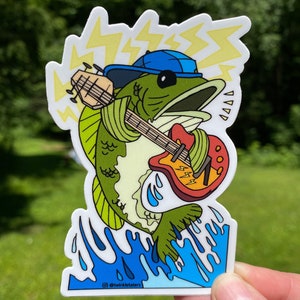May include: A green bass fish wearing a blue baseball cap and playing a red, yellow, and black electric guitar. The fish is jumping out of blue water with yellow lightning bolts behind it. The sticker has the text "@twinkletaters" at the bottom.