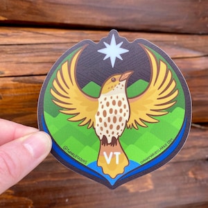 May include: A sticker featuring a stylized bird with outstretched yellow wings, a speckled body, and a blue and yellow tail. The bird is set against a green and black background with a star and the letters "VT". The sticker has the text "@twinkletaters" and "dianamarieclarke.com".