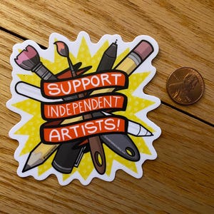 Puede incluir: Una pegatina con un dise&ntilde;o de explosi&oacute;n amarilla que presenta &uacute;tiles de arte como un pincel, un l&aacute;piz y un bol&iacute;grafo. La pegatina tiene una pancarta roja con el texto "SUPPORT INDEPENDENT ARTISTS!". Una moneda de un centavo est&aacute; al lado de la pegatina.