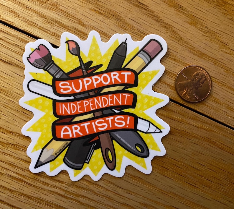 Puede incluir: Una pegatina con un dise&ntilde;o de explosi&oacute;n amarilla que presenta &uacute;tiles de arte como un pincel, un l&aacute;piz y un bol&iacute;grafo. La pegatina tiene una pancarta roja con el texto "SUPPORT INDEPENDENT ARTISTS!". Una moneda de un centavo est&aacute; al lado de la pegatina.
