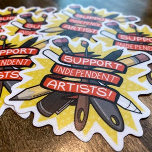 Puede incluir: Una colecci&oacute;n de pegatinas con un dise&ntilde;o de estrella amarilla y el texto "SUPPORT INDEPENDENT ARTISTS!" en pancartas rojas. Las pegatinas presentan ilustraciones de materiales de arte como pinceles, l&aacute;pices y cuchillos.