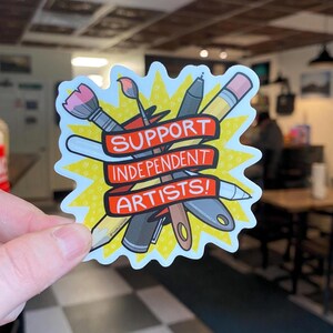 Puede incluir: Una pegatina con un dise&ntilde;o de explosi&oacute;n amarilla que presenta &uacute;tiles de arte como un pincel, un bol&iacute;grafo, un l&aacute;piz y una esp&aacute;tula. Una pancarta roja en el centro dice "SUPPORT INDEPENDENT ARTISTS!"
