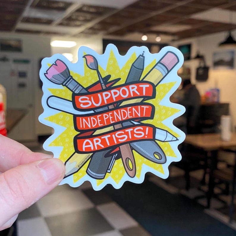 Puede incluir: Una pegatina con un dise&ntilde;o de explosi&oacute;n amarilla que presenta &uacute;tiles de arte como un pincel, un bol&iacute;grafo, un l&aacute;piz y una esp&aacute;tula. Una pancarta roja en el centro dice "SUPPORT INDEPENDENT ARTISTS!"