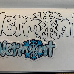May include: A blue and white sticker with the word "Vermont" in bold, black letters. The sticker has a snowflake pattern and a large blue snowflake in the center.