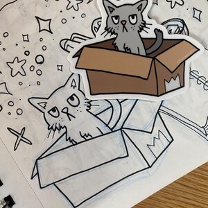 K&ouml;nnte beinhalten: Zwei Comic-Katzen mit m&uuml;rrischen Gesichtsausdr&uuml;cken sind in Kisten gezeichnet. Die obere Katze ist grau und die untere Katze ist wei&szlig;. Beide Katzen schauen nach links. Die Kisten sind braun mit einer wei&szlig;en Krone an der Seite.