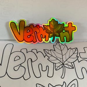 May include: A colorful sticker with the word "Vermont" in rainbow letters and a green maple leaf. The sticker is placed on a page of a coloring book with a black and white outline of the word "Vermont" and a maple leaf.