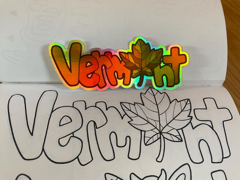 May include: A colorful sticker with the word "Vermont" in rainbow letters and a green maple leaf. The sticker is placed on a page of a coloring book with a black and white outline of the word "Vermont" and a maple leaf.
