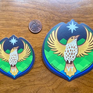 Puede incluir: Dos pegatinas con un dise&ntilde;o de p&aacute;jaro con alas extendidas, una estrella y las letras "VT". El p&aacute;jaro tiene alas amarillas y un cuerpo blanco y marr&oacute;n. Las pegatinas est&aacute;n sobre una superficie de madera, con una moneda de un centavo para la escala.