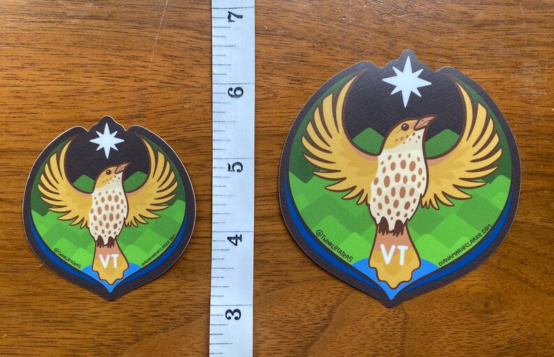 Puede incluir: Dos pegatinas con un dise&ntilde;o de p&aacute;jaro con las alas extendidas, una estrella y las letras "VT". Las pegatinas est&aacute;n sobre una superficie de madera, con una regla que muestra que la pegatina m&aacute;s grande mide aproximadamente 10 cm de alto.