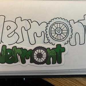 May include: A green and black sticker with the word "Vermont" and a bicycle wheel design. The sticker is on a white notebook with the word "Vermont" written in black.