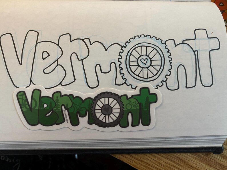May include: A green and black sticker with the word "Vermont" and a bicycle wheel design. The sticker is on a white notebook with the word "Vermont" written in black.
