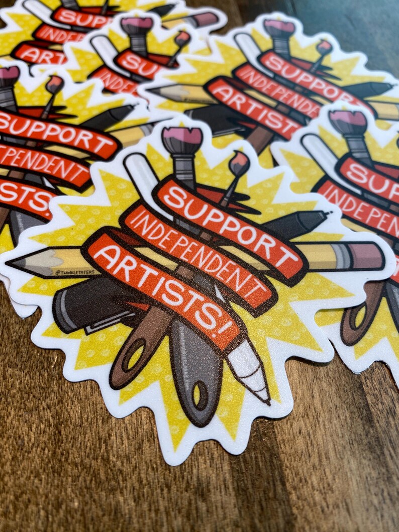 Puede incluir: Una colecci&oacute;n de pegatinas con un dise&ntilde;o de estrella amarilla. Cada pegatina presenta el texto "SUPPORT INDEPENDENT ARTISTS!" en rojo sobre una pancarta blanca, junto con ilustraciones de &uacute;tiles de arte como l&aacute;pices, pinceles y bol&iacute;grafos.