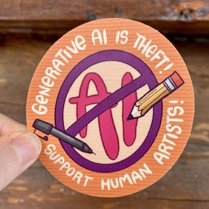 May include: Round sticker with the text "Generative AI is Theft! Support Human Artists!" in white, surrounding a purple circle with a crossed-out pencil and the letters "AI" in pink. A black pen is also visible.