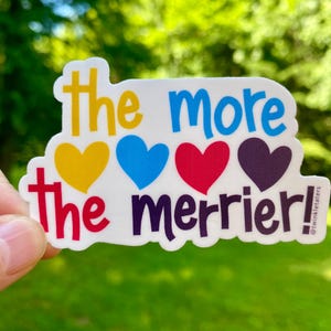 May include: A white sticker with the phrase "the more the merrier!" in yellow, blue, red, and purple. The design includes four colorful hearts. The sticker is held against a blurred green background.